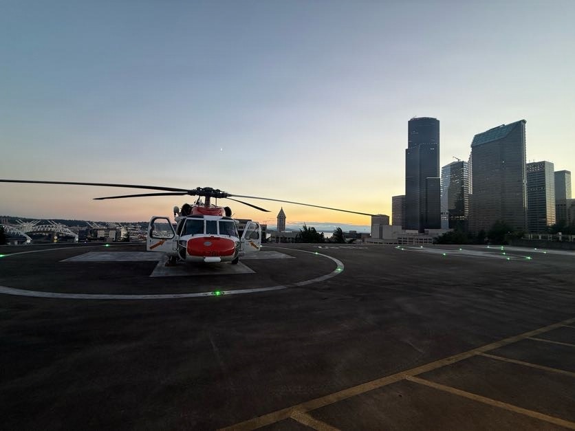 A red and white helicopter, consistent with U.S. Coast Guard livery, is parked on a rooftop helipad with green perimeter lights illuminated. The scene is captured during twilight/dusk with a city skyline visible in the background, featuring tall glass skyscrapers consistent with Seattle, Washington. The helipad appears to be atop a building, with a parking structure visible in the foreground.
