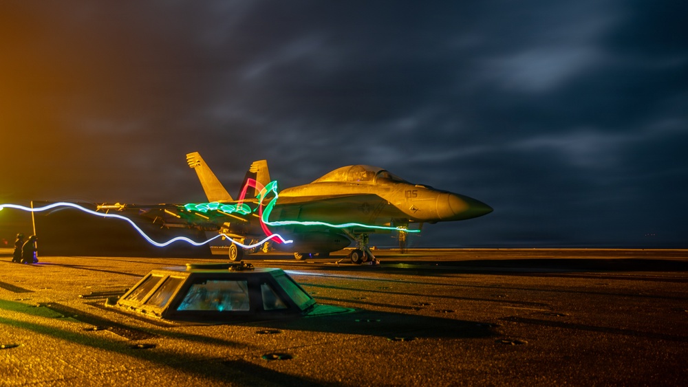 A long-exposure night photograph of a military jet fighter, likely an F/A-18 Hornet or Super Hornet, parked on what appears to be an aircraft carrier flight deck. Colorful light trails in blue, green, and red are visible around the aircraft, created by the movement of crew members carrying light wands or flashlights during the long exposure. The dramatic dark sky with clouds and warm deck lighting create a striking visual composition.