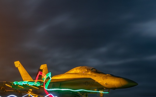 A long-exposure night photograph of a military jet fighter, likely an F/A-18 Hornet or Super Hornet, parked on what appears to be an aircraft carrier flight deck. Colorful light trails in blue, green, and red are visible around the aircraft, created by the movement of crew members carrying light wands or flashlights during the long exposure. The dramatic dark sky with clouds and warm deck lighting create a striking visual composition.