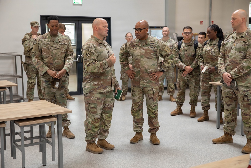 A group of U.S. Army soldiers in Operational Camouflage Pattern (OCP) uniforms are gathered inside what appears to be a modern dining facility or briefing room. Two soldiers in the foreground appear to be engaged in a discussion or briefing, with one gesturing while speaking. The setting suggests an official visit, inspection, or orientation at a military installation.