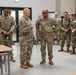 A group of U.S. Army soldiers in Operational Camouflage Pattern (OCP) uniforms are gathered inside what appears to be a modern dining facility or briefing room. Two soldiers in the foreground appear to be engaged in a discussion or briefing, with one gesturing while speaking. The setting suggests an official visit, inspection, or orientation at a military installation.