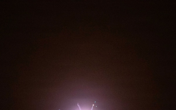 A nighttime photograph showing an aircraft or aerial object illuminated by a bright light source, possibly during takeoff, landing, or a weapons firing event. The scene is extremely dark with the subject only partially visible due to the intense light bloom. Small figures or structures are faintly visible along the bottom edge of the frame.