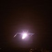 A nighttime photograph showing an aircraft or aerial object illuminated by a bright light source, possibly during takeoff, landing, or a weapons firing event. The scene is extremely dark with the subject only partially visible due to the intense light bloom. Small figures or structures are faintly visible along the bottom edge of the frame.