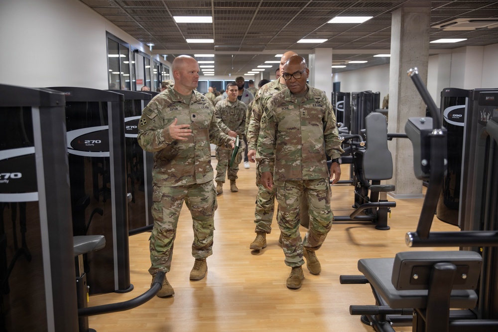 Two U.S. Army soldiers in OCP (Operational Camouflage Pattern) uniforms walk through a modern military fitness facility, appearing to conduct a tour or inspection. The soldier on the left is speaking and gesturing, while the soldier on the right, who appears to be of higher rank based on visible insignia, listens attentively. Additional uniformed personnel are visible in the background.
