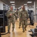 Two U.S. Army soldiers in OCP (Operational Camouflage Pattern) uniforms walk through a modern military fitness facility, appearing to conduct a tour or inspection. The soldier on the left is speaking and gesturing, while the soldier on the right, who appears to be of higher rank based on visible insignia, listens attentively. Additional uniformed personnel are visible in the background.