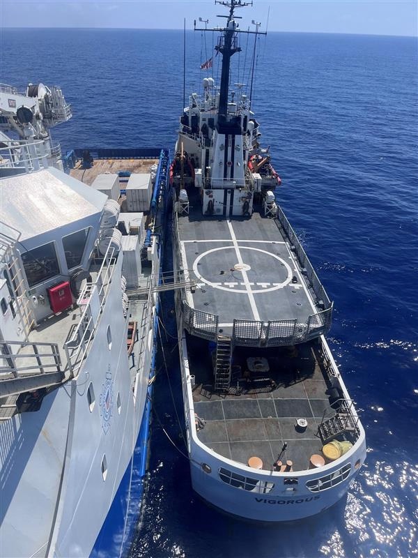 An aerial view shows two vessels moored alongside each other in open blue ocean waters. The vessel on the right, named 'VIGOROUS,' features a prominent helicopter landing pad (helipad) on its aft deck and a tall communications/navigation mast. The vessel on the left appears to be a larger ship, possibly a coast guard or law enforcement vessel, with blue hull markings and deck equipment.