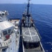 An aerial view shows two vessels moored alongside each other in open blue ocean waters. The vessel on the right, named 'VIGOROUS,' features a prominent helicopter landing pad (helipad) on its aft deck and a tall communications/navigation mast. The vessel on the left appears to be a larger ship, possibly a coast guard or law enforcement vessel, with blue hull markings and deck equipment.