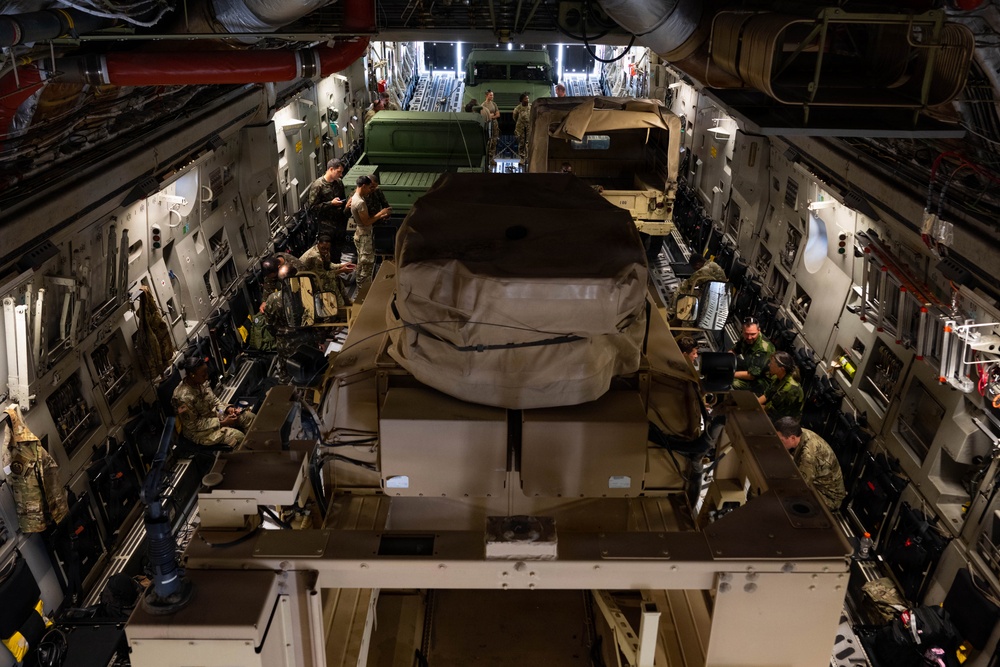 The interior of a large military cargo aircraft, likely a C-17 Globemaster III, is shown loaded with military vehicles and personnel. Multiple soldiers in camouflage uniforms are positioned along the sides of the cargo bay, appearing to conduct checks or documentation. The aircraft's cargo hold is filled with covered military vehicles secured to the floor with tie-down straps.