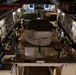 The interior of a large military cargo aircraft, likely a C-17 Globemaster III, is shown loaded with military vehicles and personnel. Multiple soldiers in camouflage uniforms are positioned along the sides of the cargo bay, appearing to conduct checks or documentation. The aircraft's cargo hold is filled with covered military vehicles secured to the floor with tie-down straps.