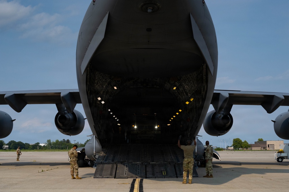 A large military cargo aircraft, consistent with a C-5 Galaxy or C-17 Globemaster, is shown with its rear cargo ramp fully lowered on a tarmac. Several military personnel in camouflage uniforms are positioned around the open cargo bay, appearing to oversee loading or unloading operations. The interior cargo hold is illuminated with yellow guide lights along the floor and ceiling.