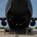 A large military cargo aircraft, consistent with a C-5 Galaxy or C-17 Globemaster, is shown with its rear cargo ramp fully lowered on a tarmac. Several military personnel in camouflage uniforms are positioned around the open cargo bay, appearing to oversee loading or unloading operations. The interior cargo hold is illuminated with yellow guide lights along the floor and ceiling.