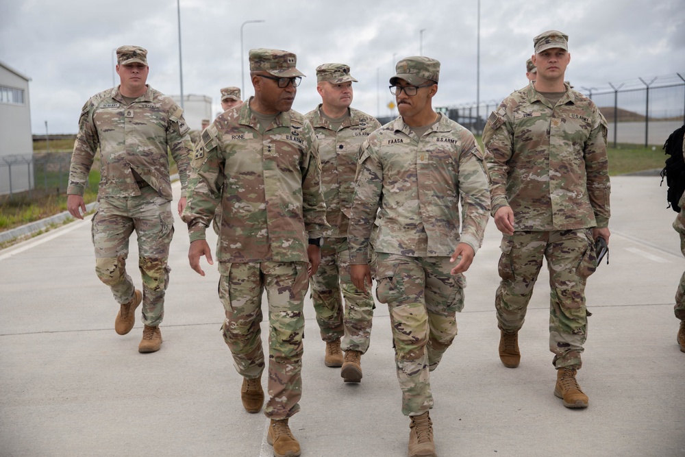 A group of U.S. Army soldiers in OCP (Operational Camouflage Pattern) uniforms are walking together on a paved surface at what appears to be a military installation or base. The group includes personnel of varying ranks, with name tapes visible on several uniforms including 'RONE,' 'FAASA,' and 'LONG.' The setting appears to be an outdoor facility with fencing and infrastructure visible in the background.