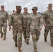A group of U.S. Army soldiers in OCP (Operational Camouflage Pattern) uniforms are walking together on a paved surface at what appears to be a military installation or base. The group includes personnel of varying ranks, with name tapes visible on several uniforms including 'RONE,' 'FAASA,' and 'LONG.' The setting appears to be an outdoor facility with fencing and infrastructure visible in the background.