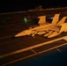 A U.S. Navy EA-18G Growler electronic warfare aircraft, marked with bureau number 506 and squadron designation VAQ-141, is positioned on an aircraft carrier flight deck at night. The aircraft is loaded with external stores including what appear to be fuel tanks and electronic warfare pods. The scene is illuminated by artificial deck lighting, with flight deck personnel visible in the background.