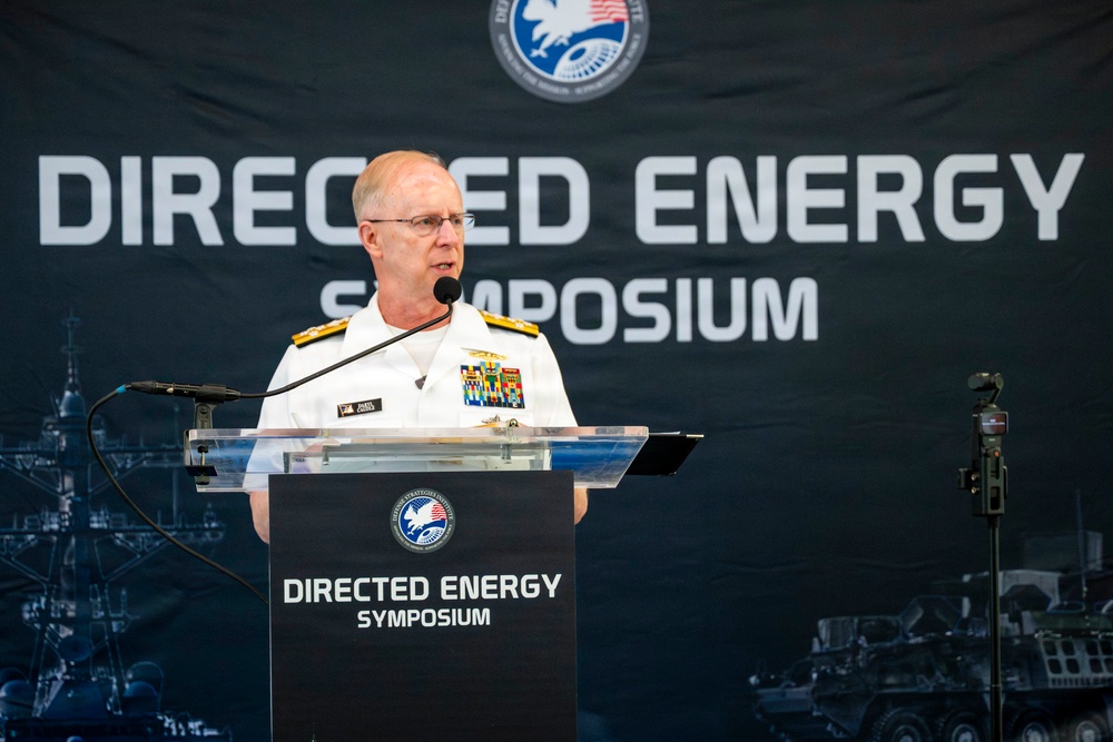 A senior U.S. Navy officer in white dress uniform is speaking at a podium during the Directed Energy Symposium. The officer displays multiple ribbons and rank insignia consistent with flag officer grade. The event appears to be a formal military-academic or defense industry conference.