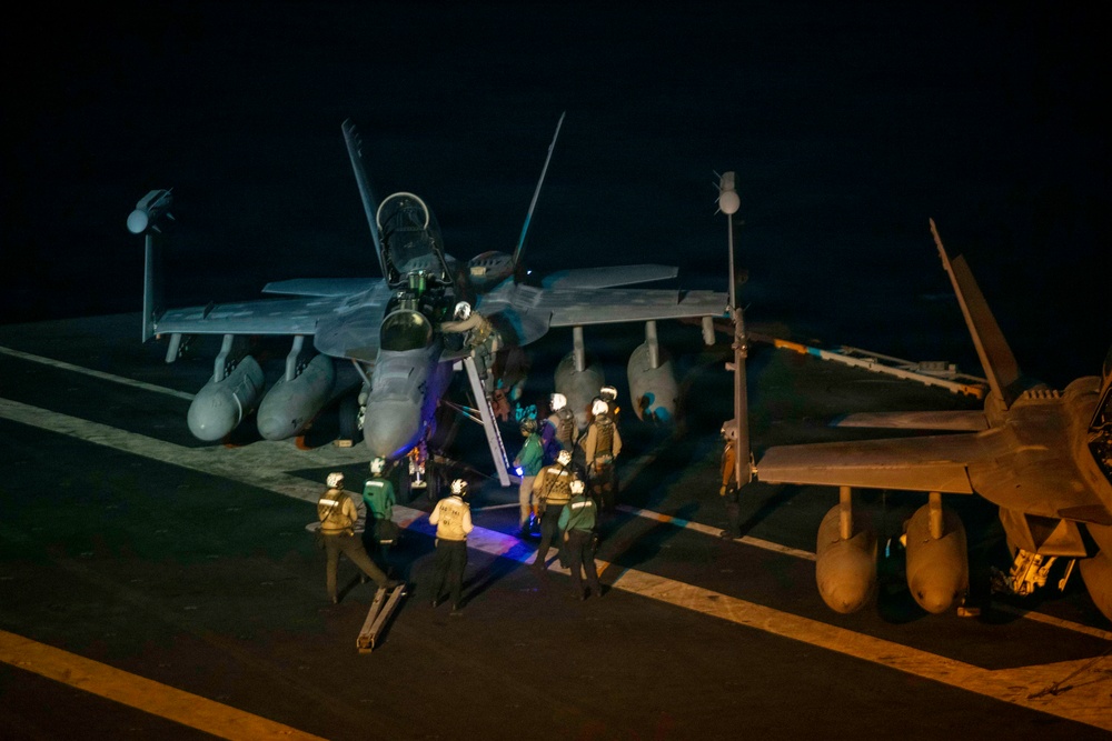A nighttime scene on what appears to be a naval aircraft carrier flight deck, showing military personnel gathered around a twin-engine jet aircraft, likely an F/A-18 Hornet or Super Hornet. Ground crew members in various colored jerseys are conducting pre-flight or post-flight operations around the aircraft. At least one person appears to be climbing into or out of the cockpit via a ladder.
