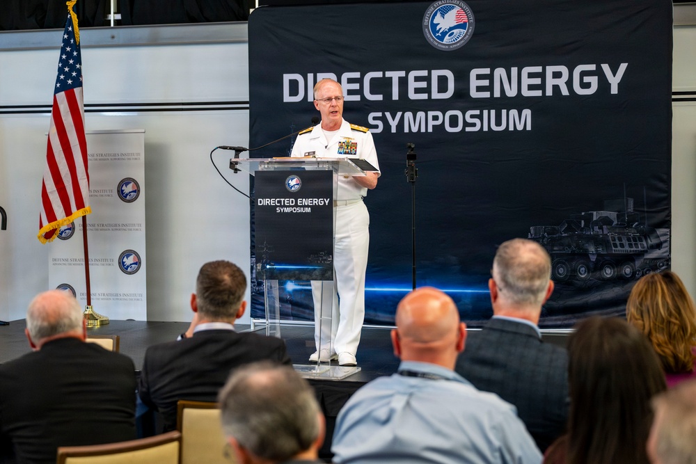 A U.S. Navy officer in white dress uniform is delivering a speech at a podium during the Directed Energy Symposium, organized by the Defense Strategies Institute. The event appears to be a professional defense industry conference with an audience of civilians and possibly military personnel seated in the foreground. The backdrop features military imagery including what appears to be armored vehicles.