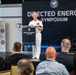 A U.S. Navy officer in white dress uniform is delivering a speech at a podium during the Directed Energy Symposium, organized by the Defense Strategies Institute. The event appears to be a professional defense industry conference with an audience of civilians and possibly military personnel seated in the foreground. The backdrop features military imagery including what appears to be armored vehicles.