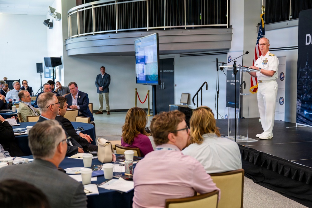 A military officer in white dress uniform is delivering a speech at a podium during what appears to be a 'Directed Energy Symposium' event. The audience is seated at round tables in a modern conference venue, attentively listening to the presentation. An American flag and a display screen with presentation slides are visible on the stage.