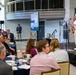 A military officer in white dress uniform is delivering a speech at a podium during what appears to be a 'Directed Energy Symposium' event. The audience is seated at round tables in a modern conference venue, attentively listening to the presentation. An American flag and a display screen with presentation slides are visible on the stage.