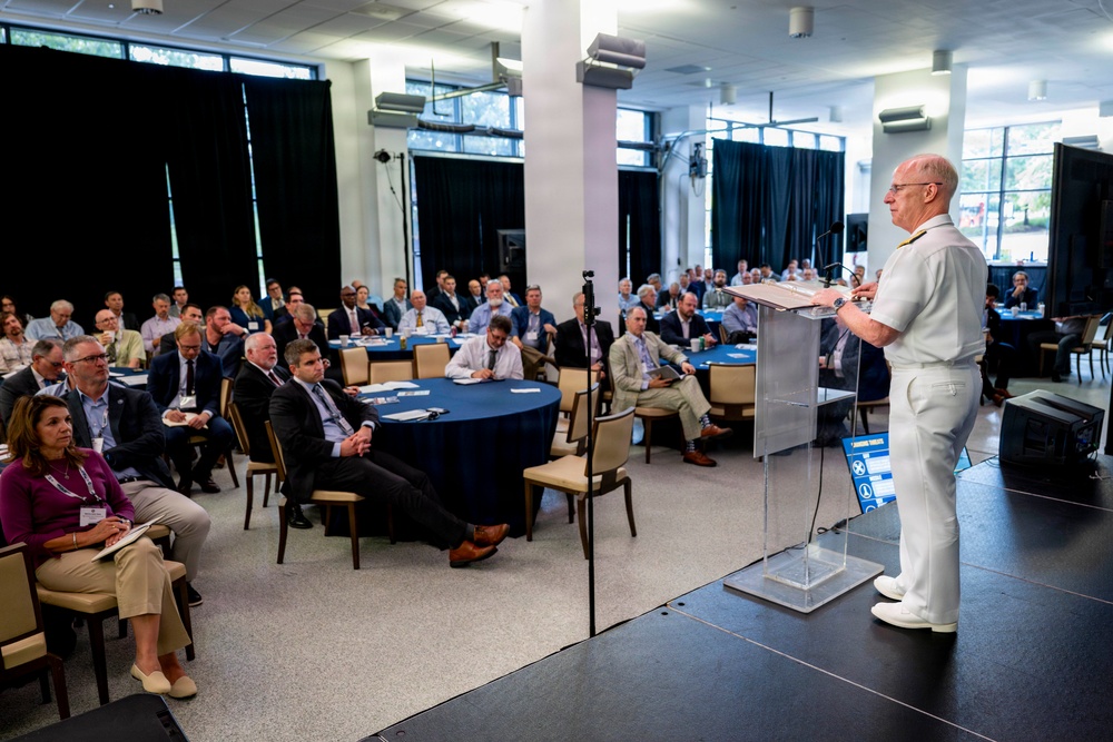 A military officer in white dress uniform is delivering a speech at a podium during a formal conference or symposium. The audience, consisting of approximately 50-60 civilians and professionals, is seated at round tables with blue tablecloths in a modern conference hall. The setting appears to be an official government or defense-related event.