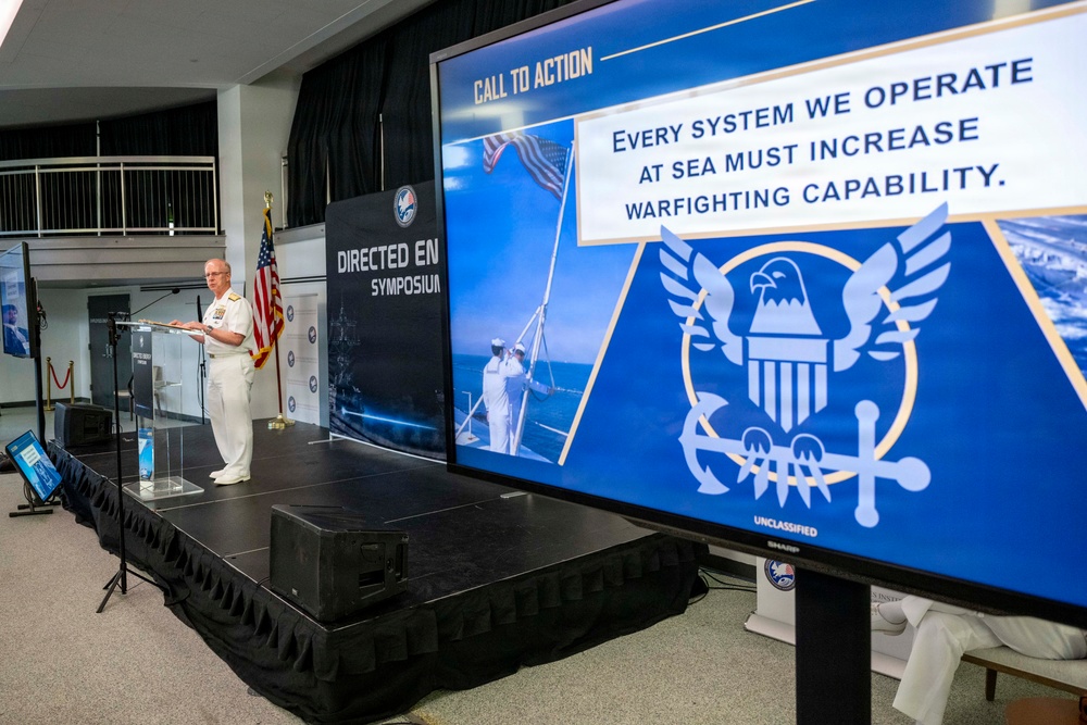 A U.S. Navy officer in white dress uniform delivers a speech at a podium during a 'Directed Energy Symposium.' A large display screen shows a 'Call to Action' slide reading 'Every System We Operate At Sea Must Increase Warfighting Capability,' marked 'UNCLASSIFIED,' featuring a Navy emblem and sailors raising a flag. The event appears to be a formal military conference or briefing.