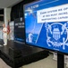 A U.S. Navy officer in white dress uniform delivers a speech at a podium during a 'Directed Energy Symposium.' A large display screen shows a 'Call to Action' slide reading 'Every System We Operate At Sea Must Increase Warfighting Capability,' marked 'UNCLASSIFIED,' featuring a Navy emblem and sailors raising a flag. The event appears to be a formal military conference or briefing.