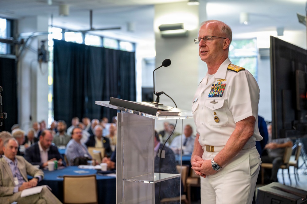 A senior U.S. Navy officer in white dress uniform stands at a clear acrylic podium delivering a speech to a seated audience. The officer displays multiple rows of service ribbons, gold shoulder boards indicating flag officer rank, and various qualification badges. The setting appears to be a formal conference or symposium held in a well-lit indoor venue.