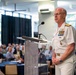 A senior U.S. Navy officer in white dress uniform stands at a clear acrylic podium delivering a speech to a seated audience. The officer displays multiple rows of service ribbons, gold shoulder boards indicating flag officer rank, and various qualification badges. The setting appears to be a formal conference or symposium held in a well-lit indoor venue.