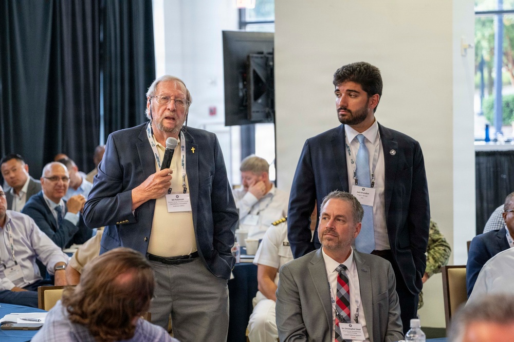 A formal conference or symposium setting where an older gentleman in a navy blazer is standing and speaking into a microphone while wearing a name badge and lanyard. He is flanked by a younger man in a dark suit also standing, while a third man in a gray suit remains seated. The room is filled with attendees seated at tables, including individuals in what appear to be military uniforms.