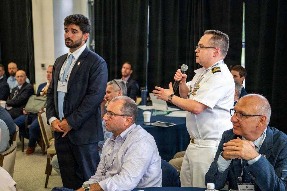 A professional conference or symposium setting where attendees are seated at round tables. A naval officer in white dress uniform is standing and speaking into a microphone, while a man in a dark suit stands nearby. The audience consists of numerous attendees wearing conference lanyards and name badges.