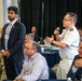 A professional conference or symposium setting where attendees are seated at round tables. A naval officer in white dress uniform is standing and speaking into a microphone, while a man in a dark suit stands nearby. The audience consists of numerous attendees wearing conference lanyards and name badges.