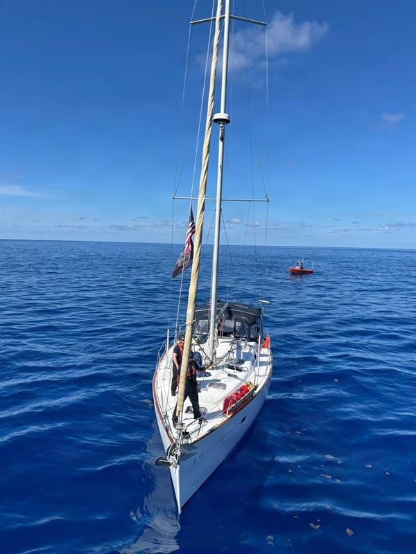 A white monohull sailboat is stationary on calm, deep blue open ocean water under clear skies. One person wearing a life vest is visible on the foredeck, and a small red vessel — consistent with a Coast Guard or rescue boat — is visible in the background. An American flag is flying from the rigging, and the sails are furled.