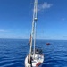 A white monohull sailboat is stationary on calm, deep blue open ocean water under clear skies. One person wearing a life vest is visible on the foredeck, and a small red vessel — consistent with a Coast Guard or rescue boat — is visible in the background. An American flag is flying from the rigging, and the sails are furled.