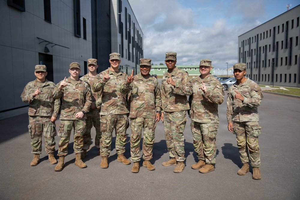 A group of nine U.S. Army soldiers in OCP (Operational Camouflage Pattern) uniforms pose for a group photo outdoors at what appears to be a military installation. Most of the soldiers are holding up one finger, suggesting a 'number one' or unit pride gesture. The setting includes modern military-style buildings in the background under a partly cloudy sky.