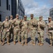A group of nine U.S. Army soldiers in OCP (Operational Camouflage Pattern) uniforms pose for a group photo outdoors at what appears to be a military installation. Most of the soldiers are holding up one finger, suggesting a 'number one' or unit pride gesture. The setting includes modern military-style buildings in the background under a partly cloudy sky.
