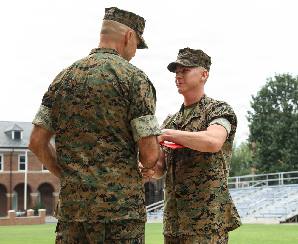 Two U.S. Marines in MARPAT woodland camouflage uniforms are engaged in a handshake during what appears to be a formal military ceremony. The Marine on the right is holding a red presentation folder or award case while shaking hands with the senior Marine whose back is to the camera. The setting appears to be an outdoor parade field or athletic field adjacent to a brick building and bleachers.