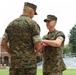 Two U.S. Marines in MARPAT woodland camouflage uniforms are engaged in a handshake during what appears to be a formal military ceremony. The Marine on the right is holding a red presentation folder or award case while shaking hands with the senior Marine whose back is to the camera. The setting appears to be an outdoor parade field or athletic field adjacent to a brick building and bleachers.
