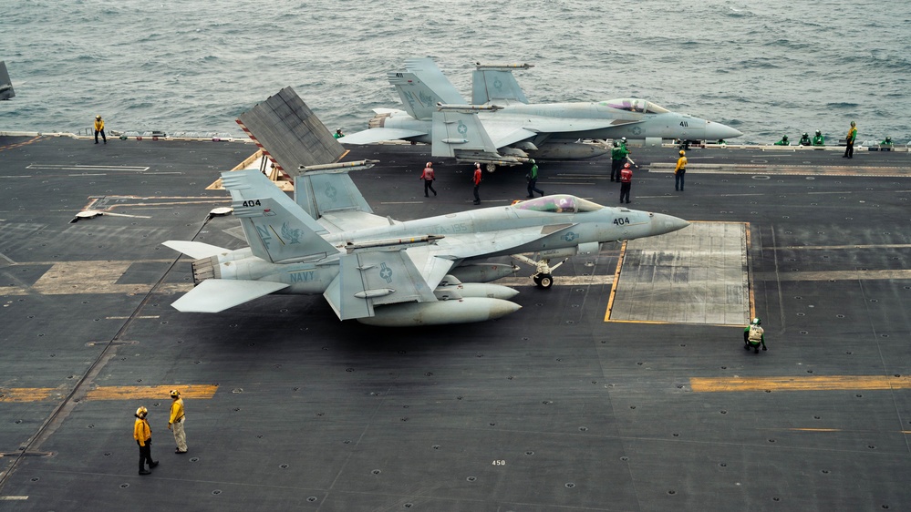 A U.S. Navy aircraft carrier flight deck shows at least two F/A-18 Hornet or Super Hornet jets positioned for operations, with bureau numbers 404 and 411 visible. Multiple flight deck crew members in color-coded jerseys (yellow, green, red) are performing their respective roles across the deck. The carrier is underway at sea, with open ocean visible in the background.
