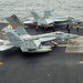 A U.S. Navy aircraft carrier flight deck shows at least two F/A-18 Hornet or Super Hornet jets positioned for operations, with bureau numbers 404 and 411 visible. Multiple flight deck crew members in color-coded jerseys (yellow, green, red) are performing their respective roles across the deck. The carrier is underway at sea, with open ocean visible in the background.