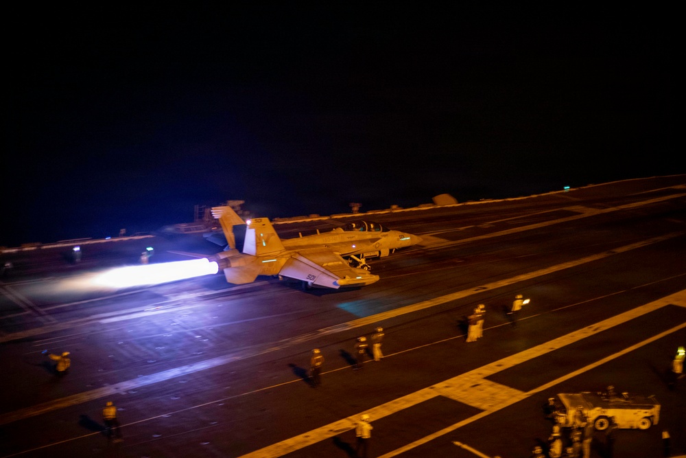 A military jet aircraft is launching from an aircraft carrier flight deck at night, with afterburners producing a bright, intense flame visible from the rear engines. Multiple flight deck personnel in colored jerseys are visible at various positions on the deck. The scene is illuminated primarily by the afterburner glow and deck lighting, creating a dramatic low-light operational photograph.