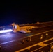 A military jet aircraft is launching from an aircraft carrier flight deck at night, with afterburners producing a bright, intense flame visible from the rear engines. Multiple flight deck personnel in colored jerseys are visible at various positions on the deck. The scene is illuminated primarily by the afterburner glow and deck lighting, creating a dramatic low-light operational photograph.
