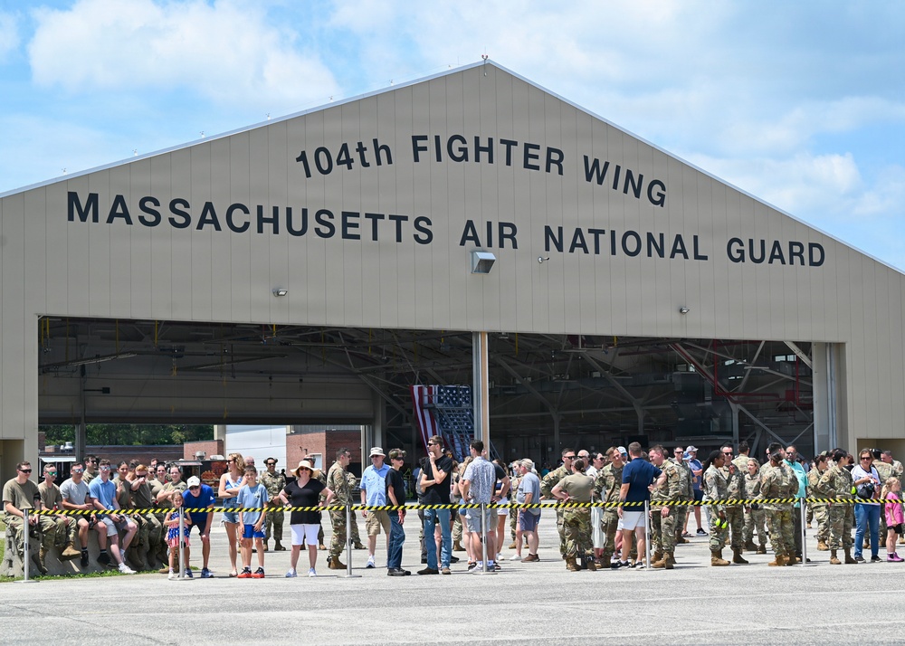 A large group of military personnel in camouflage uniforms and civilians are gathered outside a hangar belonging to the 104th Fighter Wing, Massachusetts Air National Guard. The crowd appears to be assembled for an official event, possibly a deployment sendoff, homecoming, or ceremony. Yellow and black caution tape forms a barrier line in front of the hangar.