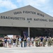 A large group of military personnel in camouflage uniforms and civilians are gathered outside a hangar belonging to the 104th Fighter Wing, Massachusetts Air National Guard. The crowd appears to be assembled for an official event, possibly a deployment sendoff, homecoming, or ceremony. Yellow and black caution tape forms a barrier line in front of the hangar.