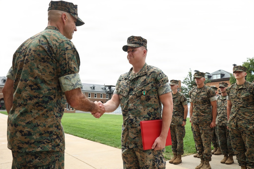 A U.S. Marine Corps award ceremony is taking place outdoors on a military installation. A senior Marine officer is shaking hands with a recipient who is wearing a medal and holding a red award folder. A formation of Marines stands at attention in the background observing the ceremony.