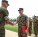 A U.S. Marine Corps award ceremony is taking place outdoors on a military installation. A senior Marine officer is shaking hands with a recipient who is wearing a medal and holding a red award folder. A formation of Marines stands at attention in the background observing the ceremony.