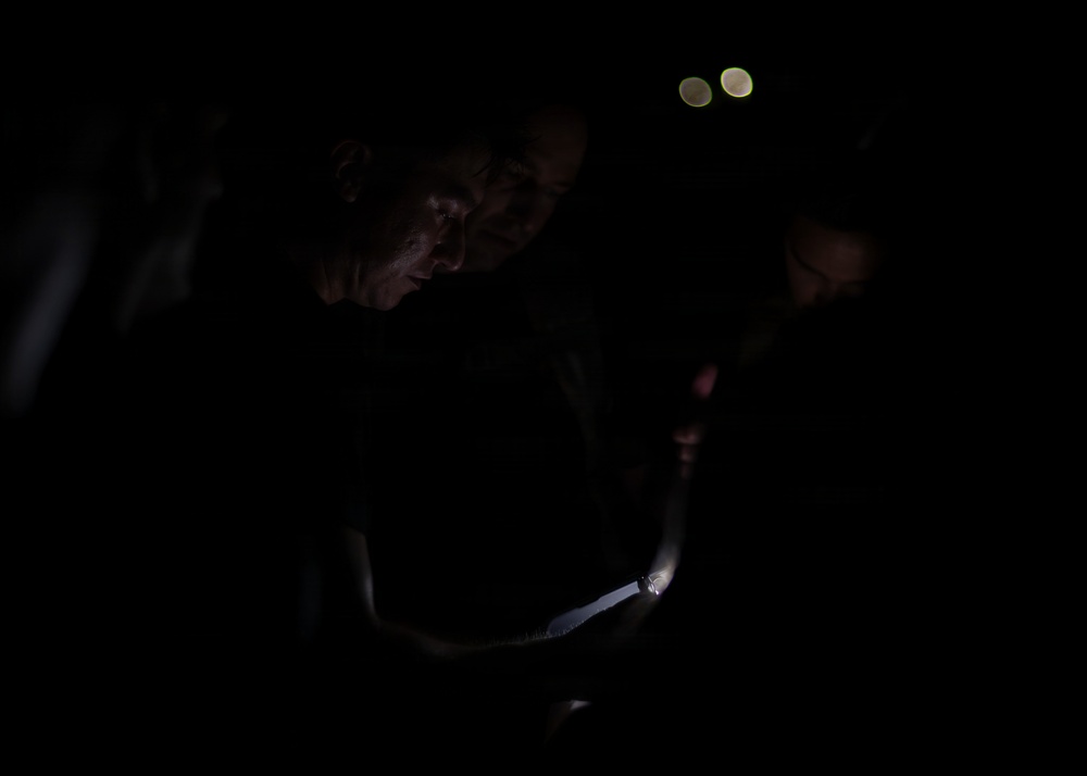 A very dark, low-light photograph showing at least two individuals illuminated primarily by a small light source in the lower center of the frame. The dominant figure on the left is leaning forward, their face partially lit from below. Two small bokeh light circles are visible in the upper background.
