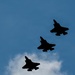 Four military jet aircraft are captured in silhouette flying in a diagonal echelon formation against a blue sky with partial cloud cover. The aircraft are rendered entirely in dark silhouette, photographed from below. Their distinctive delta-wing and twin-tail profiles are consistent with fifth-generation stealth fighter designs.