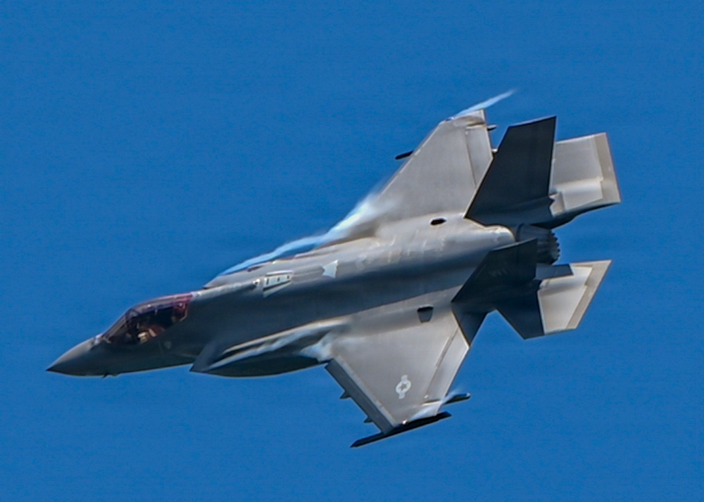A single-engine stealth multirole fighter jet, consistent with an F-35 Lightning II, is photographed in flight against a clear blue sky. The aircraft is captured in a banked maneuver, providing a clear view of its underside and tail section. The image shows fine detail of the aircraft's stealth-optimized airframe, control surfaces, and exhaust nozzle.