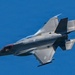A single-engine stealth multirole fighter jet, consistent with an F-35 Lightning II, is photographed in flight against a clear blue sky. The aircraft is captured in a banked maneuver, providing a clear view of its underside and tail section. The image shows fine detail of the aircraft's stealth-optimized airframe, control surfaces, and exhaust nozzle.
